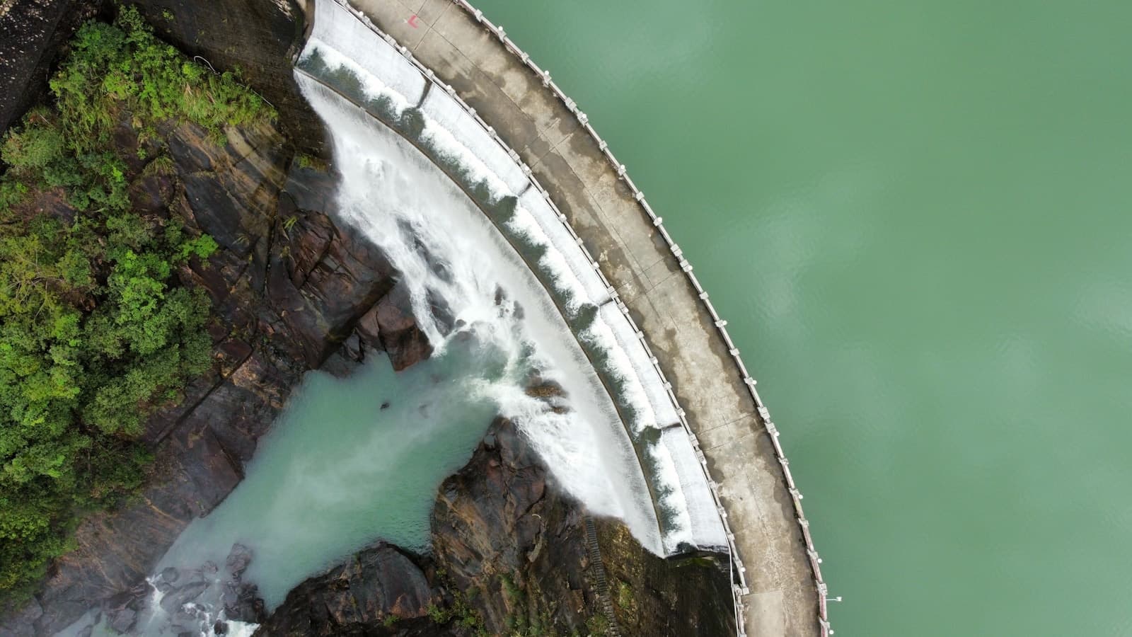 An aerial view of water flowing over the curved spillway of a dam into a rocky gorge